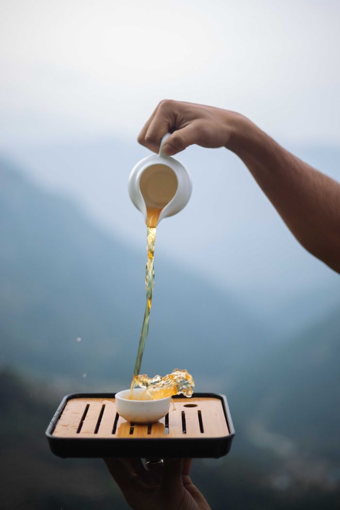 Hand gracefully pours hot tea onto a wooden tray outdoors.