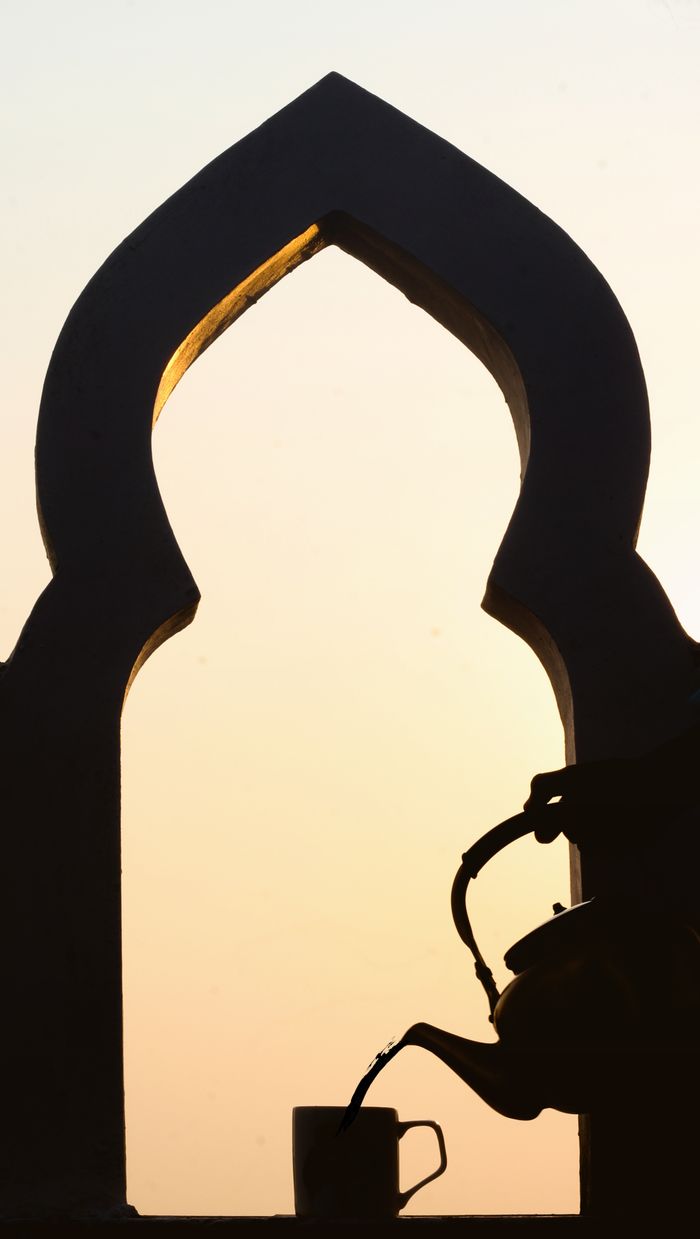 Silhouette of a teapot pouring tea into a cup by a traditional Indian arch at sunset.