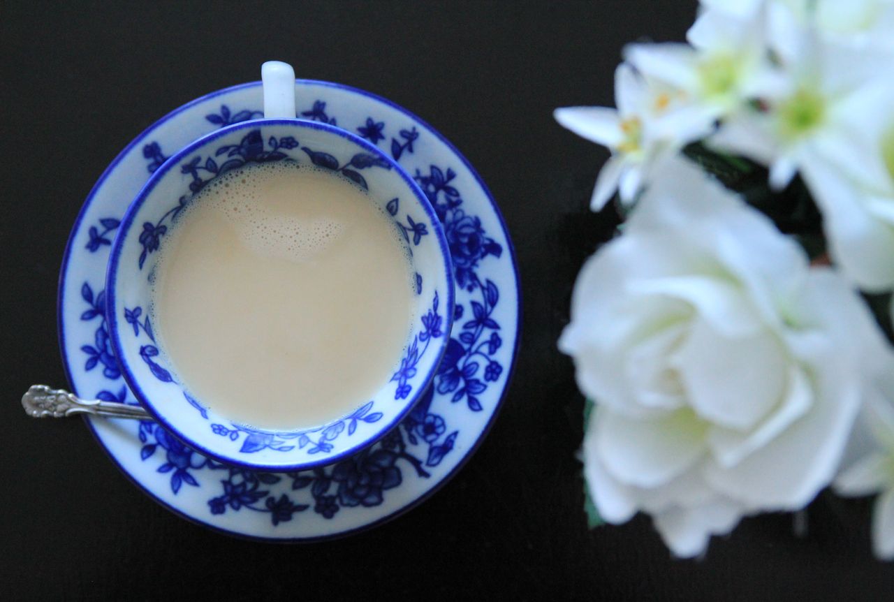 Top view of a floral tea cup with foamy beverage and white flowers on a dark surface.